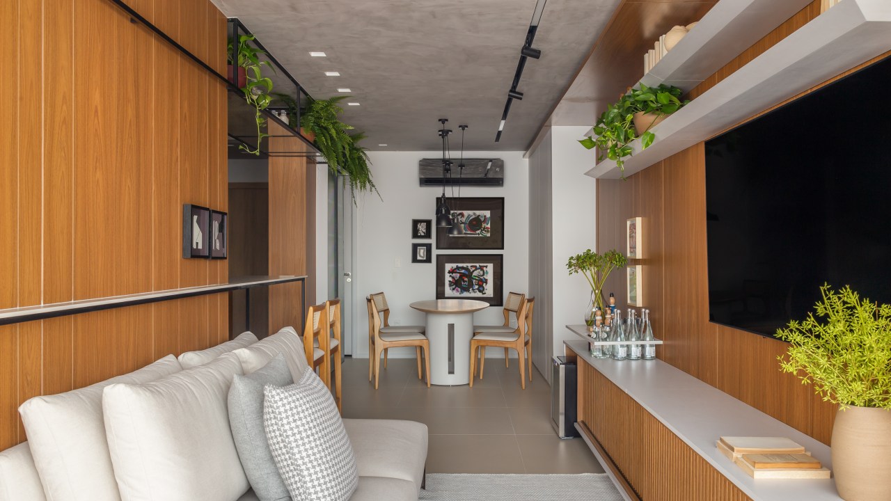 Sala de estar integrada com varanda, cozinha e estar. Sala estreita com sofá cinza e parede revestida de madeira.