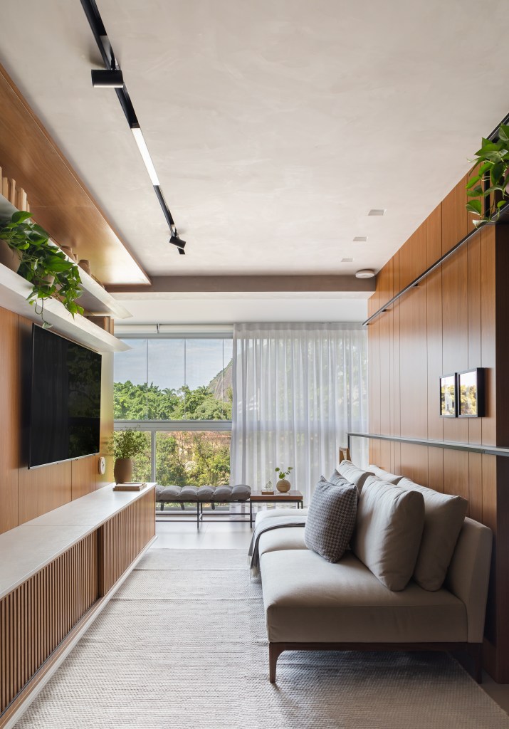 Sala de estar estreita com sofá cinza, parede revestida de madeira e painel para tv.