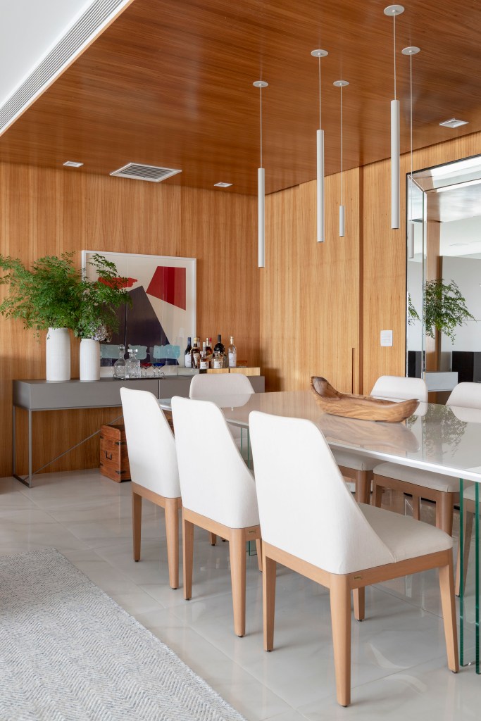 Sala de jantar com paredes revestidas de madeira, mesa de jantar branca, cadeiras estofadas brancas, aparador com quadro e vasos.