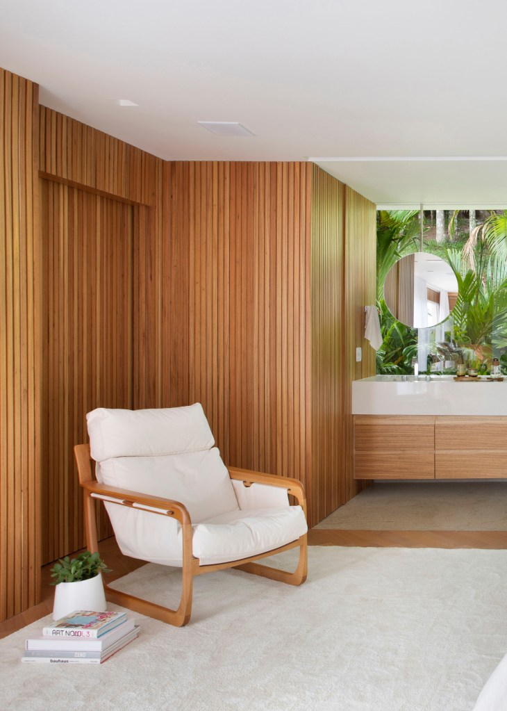 Suíte com parede revestida de madeira ripada, poltrona branca e banheiro com vista para jardim.