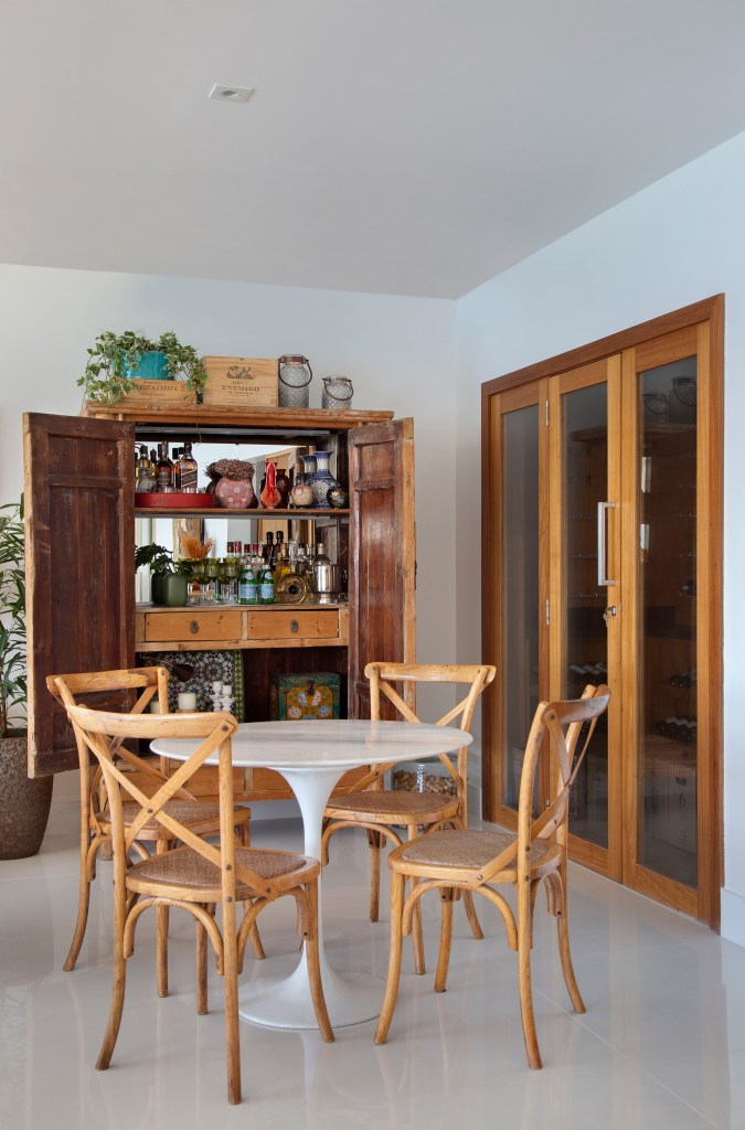 Sala de jantar pequena com mesa redonda branca e cadeiras de madeira; cristaleira de madeira.