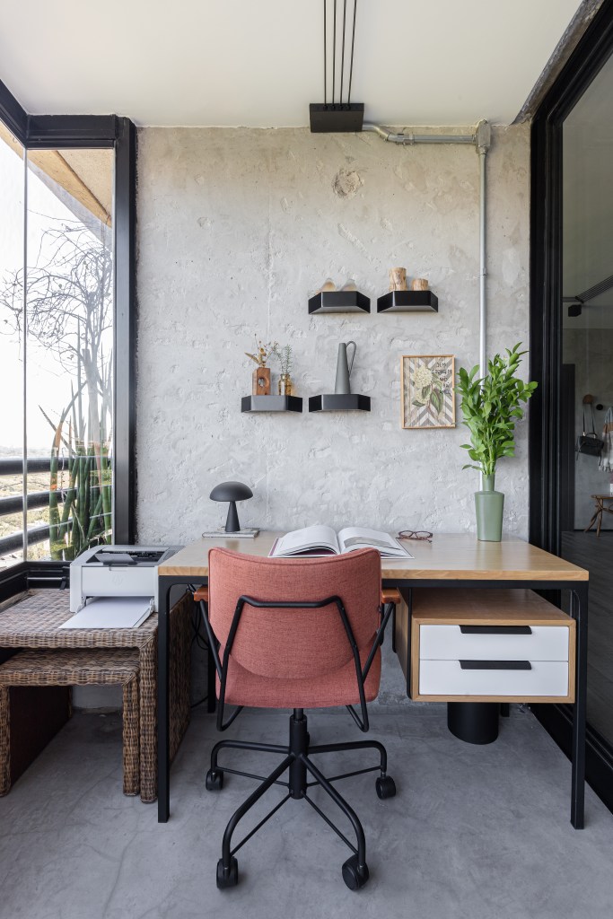 Home office na varanda com mesa L de madeira, cadeira salmão e parede de cimento queimado,