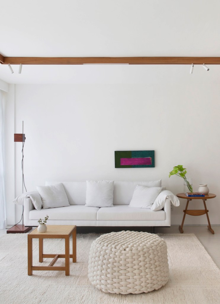 Sala minimalista com parede branca, sofá branco, pufe de macramê, tapete bege e mesa lateral de madeira.