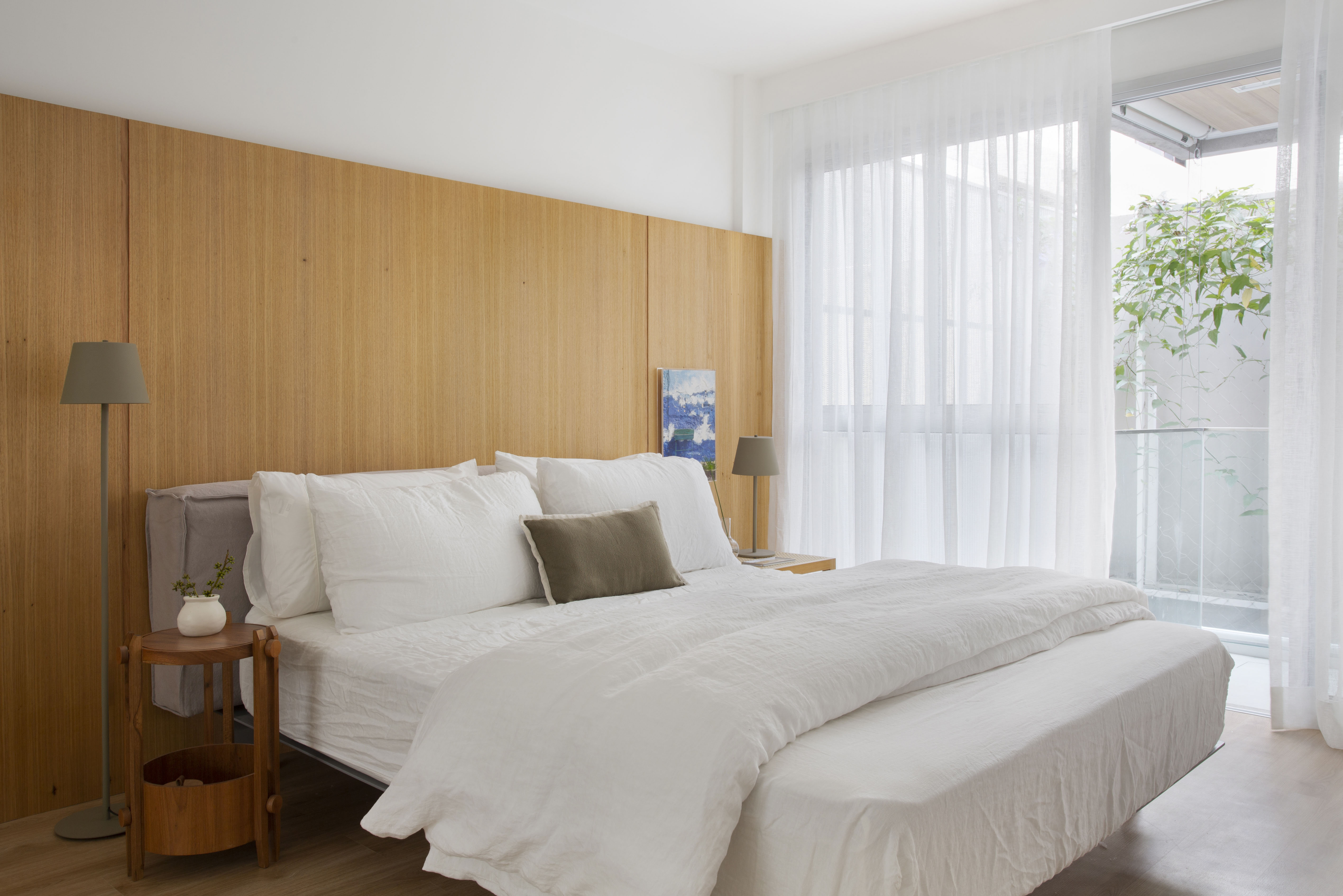 Quarto claro com cabeceira de madeira, cama de casal e cortina branca.