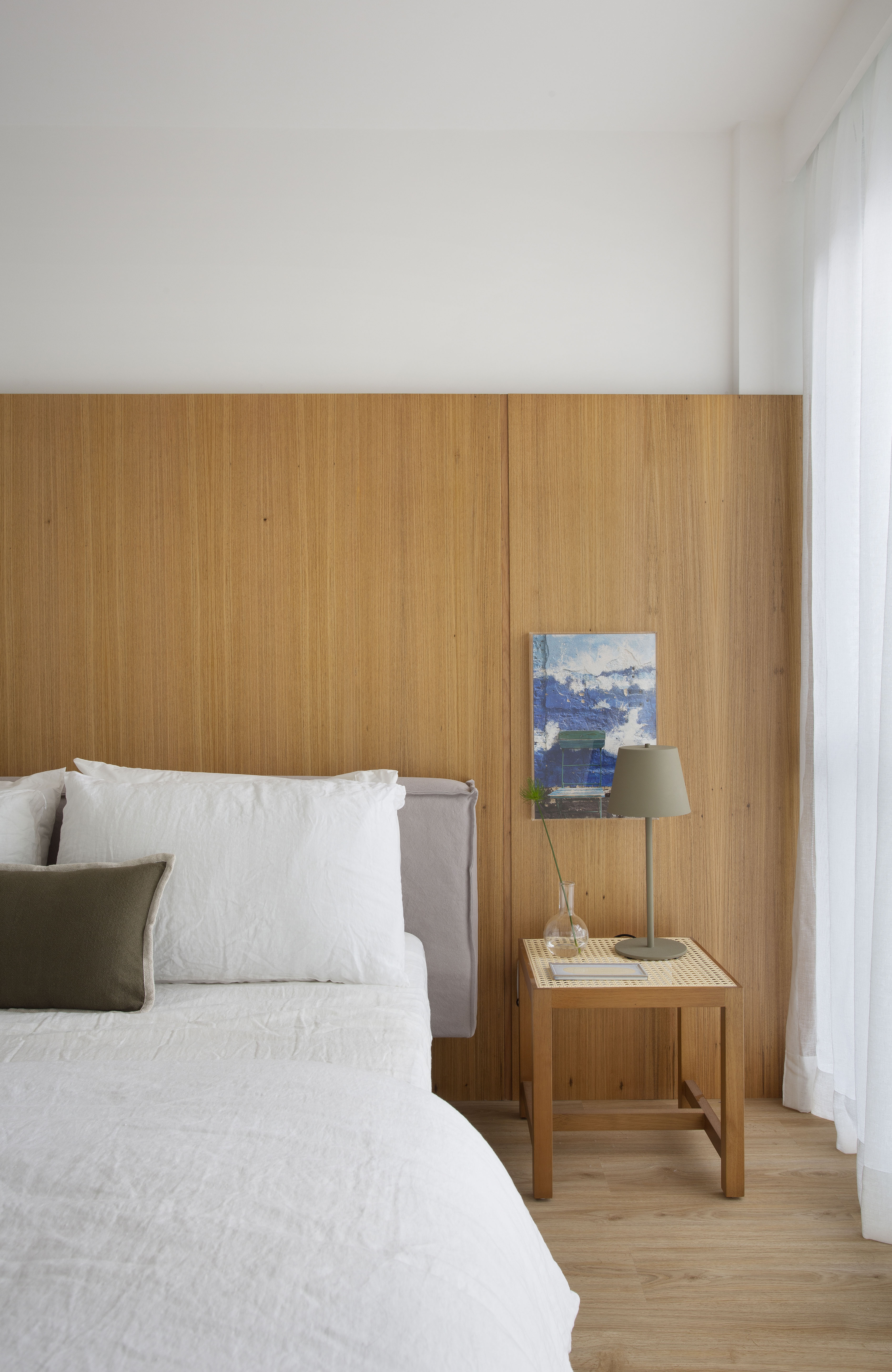 Quarto claro com cabeceira de madeira, cama de casal e cortina branca.