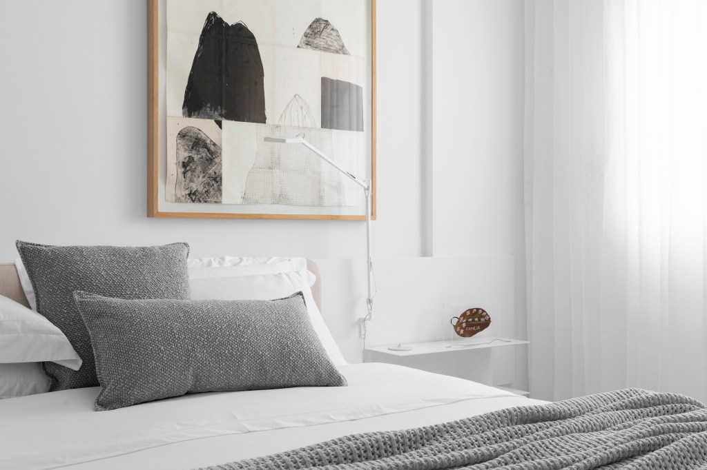 Quarto branco minimalista com cama de casal, mesinha lateral e quadro.