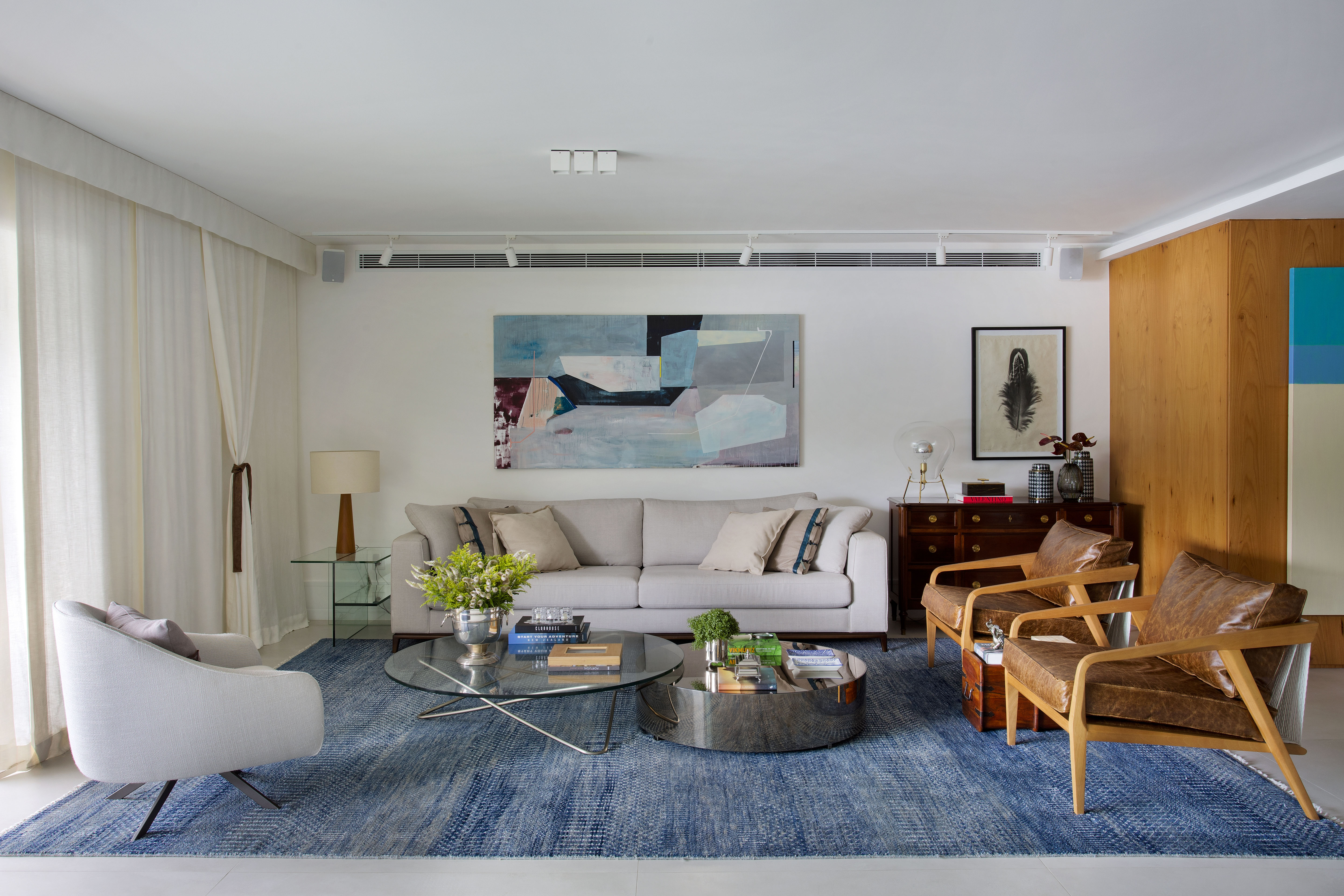 Sala de estar com sofá claro, tapete azul, quadros e cadeiras de madeira.