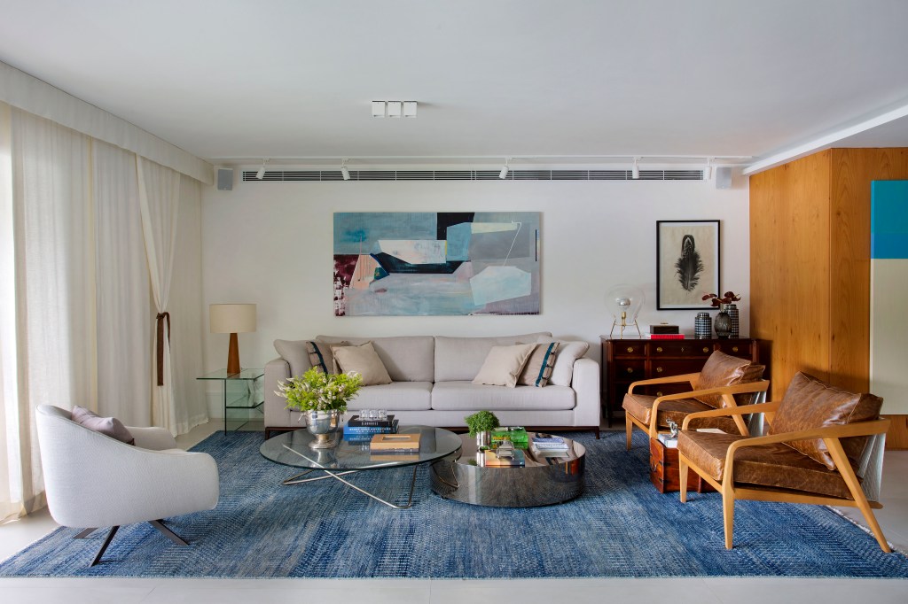 Sala de estar com sofá claro, tapete azul, quadros e cadeiras de madeira.
