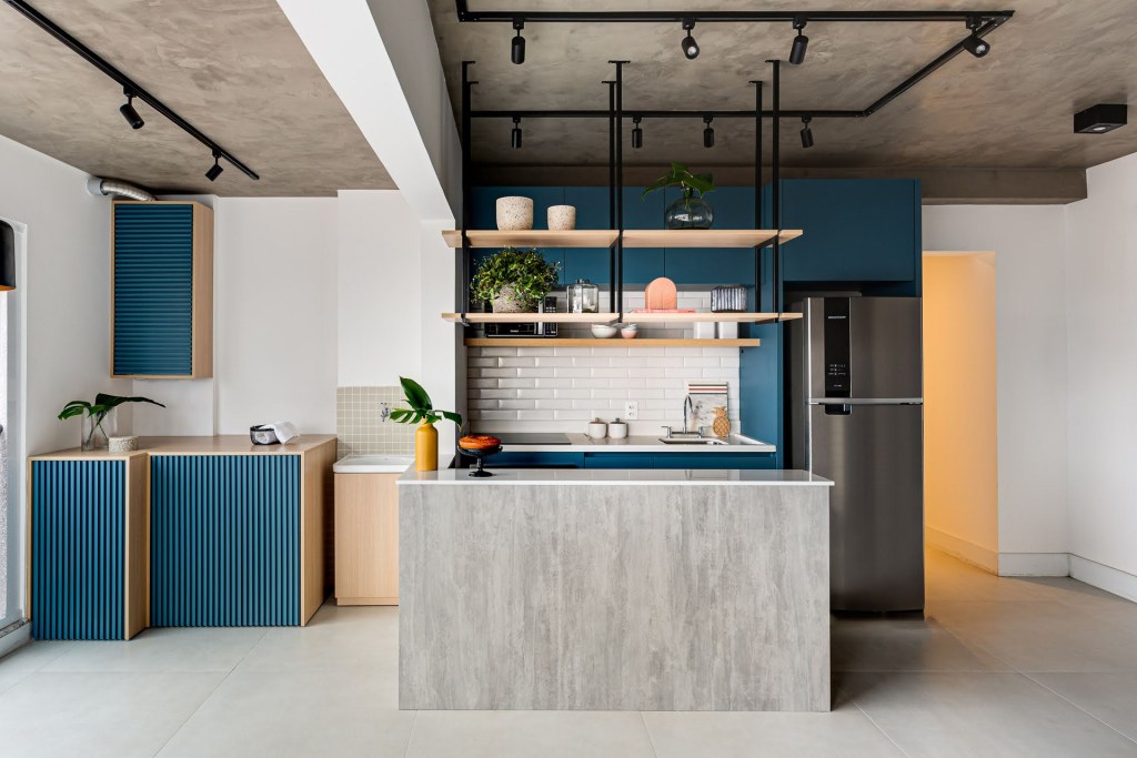 Cozinha integrada com ilha revestida de cimento queimado, prateleiras suspensas e marcenaria azul.