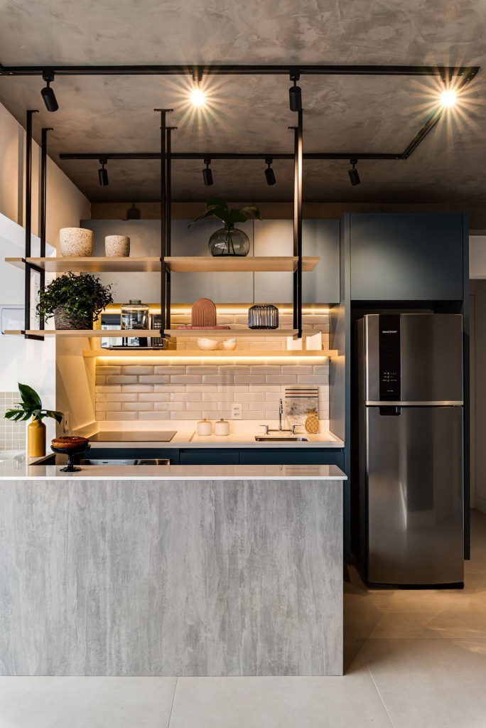 Cozinha integrada iluminada com ilha revestida de cimento queimado, prateleiras suspensas e marcenaria azul.