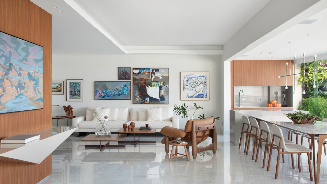Sala de estar clara com sofá branco, quadros atrás do sofá e poltrona de couro. Varanda gourmet integrada.