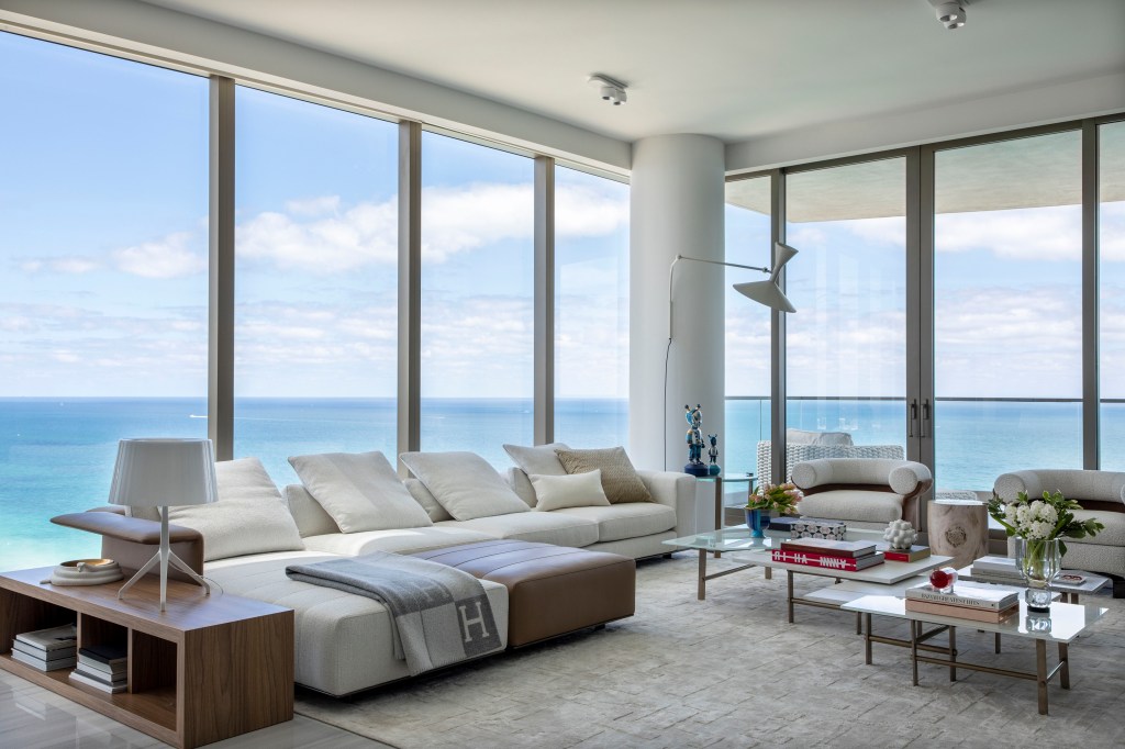 Sala de estar branca com vista para o mar, sofá e poltrona branca, luminária branca de piso e mesa de centro.