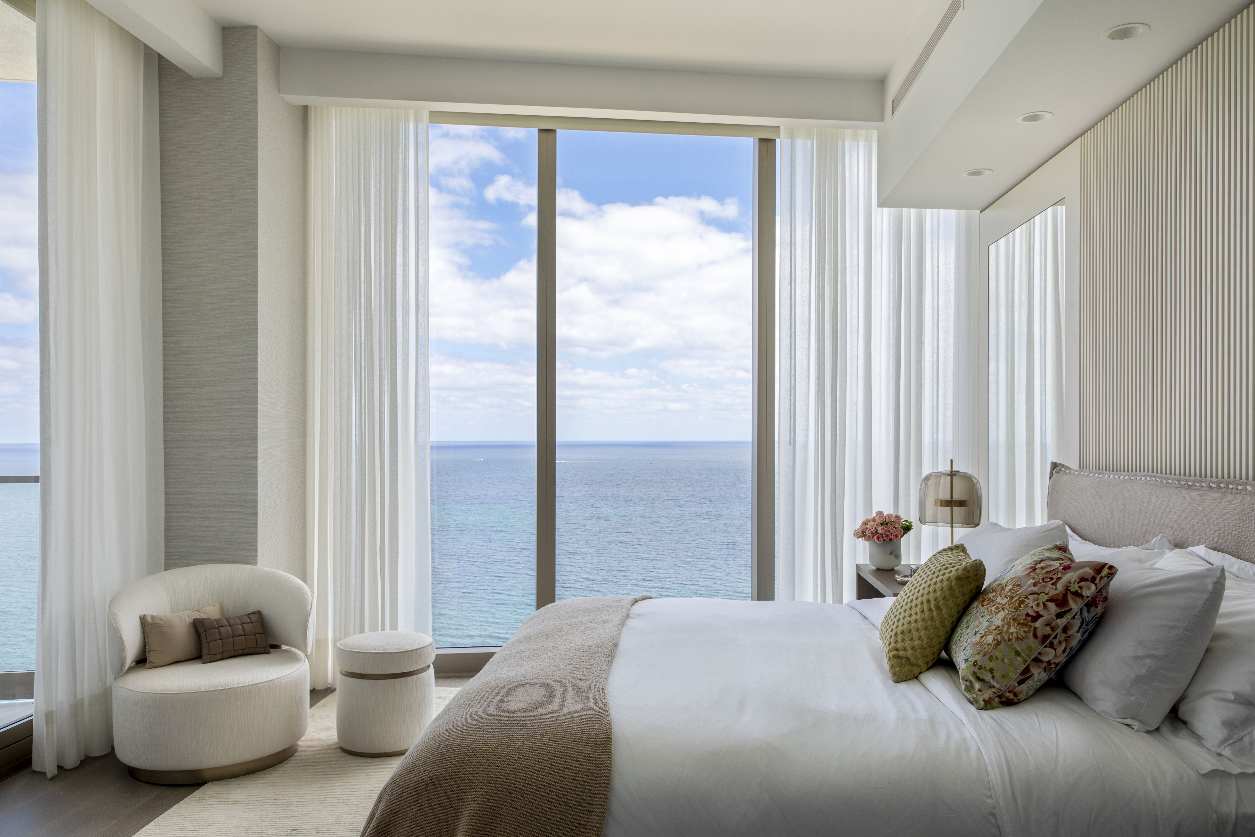 Quarto com vista para o mar, cama de casal, poltrona branca e cortina branca.