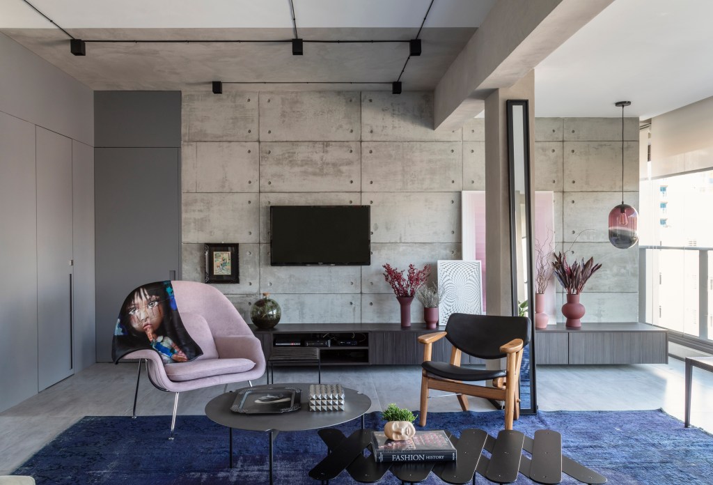 Sala de estar com paredes revestidas de cimento queimado, tapete azul, sofá cinza e poltrona rosa.