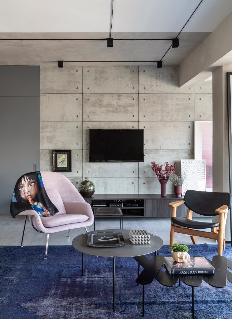 Sala de estar com paredes revestidas de cimento queimado, tapete azul, sofá cinza e poltrona rosa.