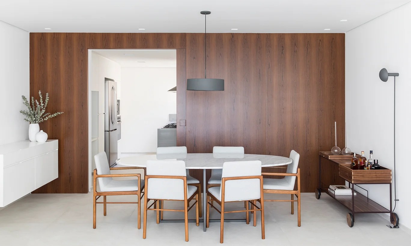 Sala de jantar com parede revestida de madeira, mesa oval branca e cadeiras brancas.