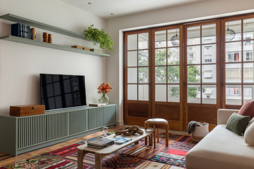Sala de tv com parede de tijolinho, rack verde claro, tapete colorido, mesa de centro em madeira e porta para varanda em madeira