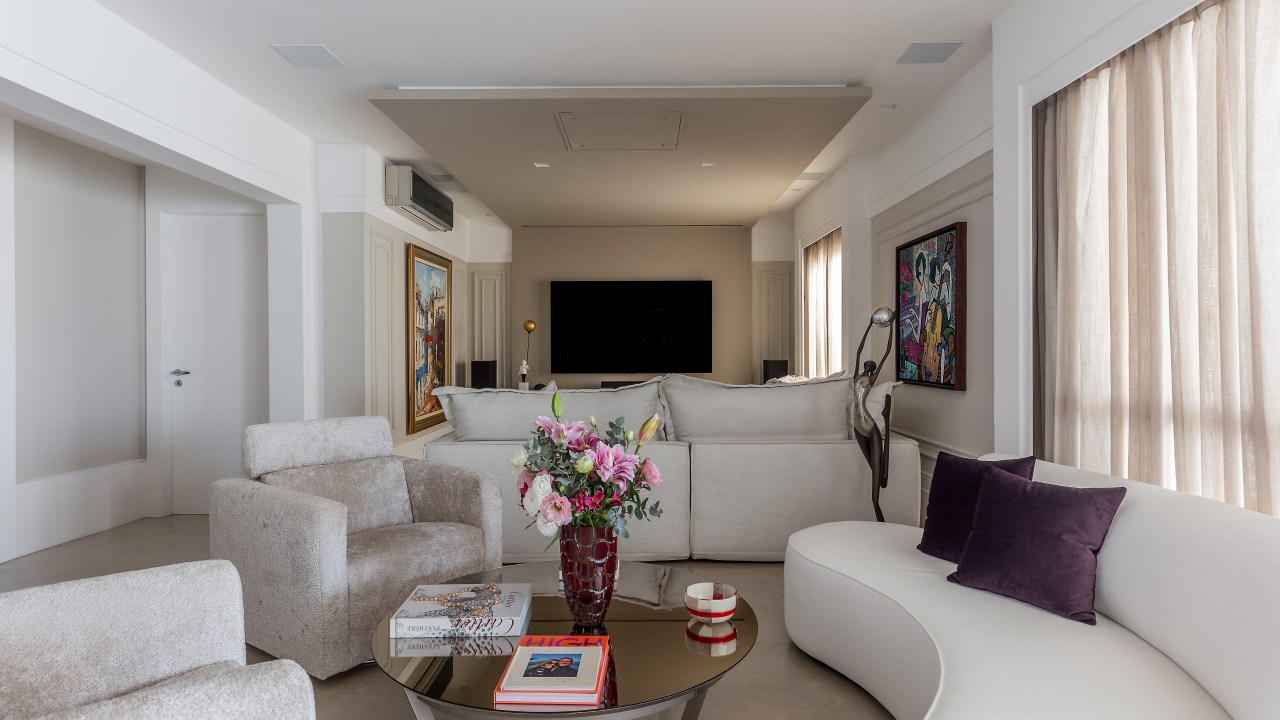 Sala de estar com sala de tv, sofás e poltronas brancas, sofá curvo e quadros nas paredes.