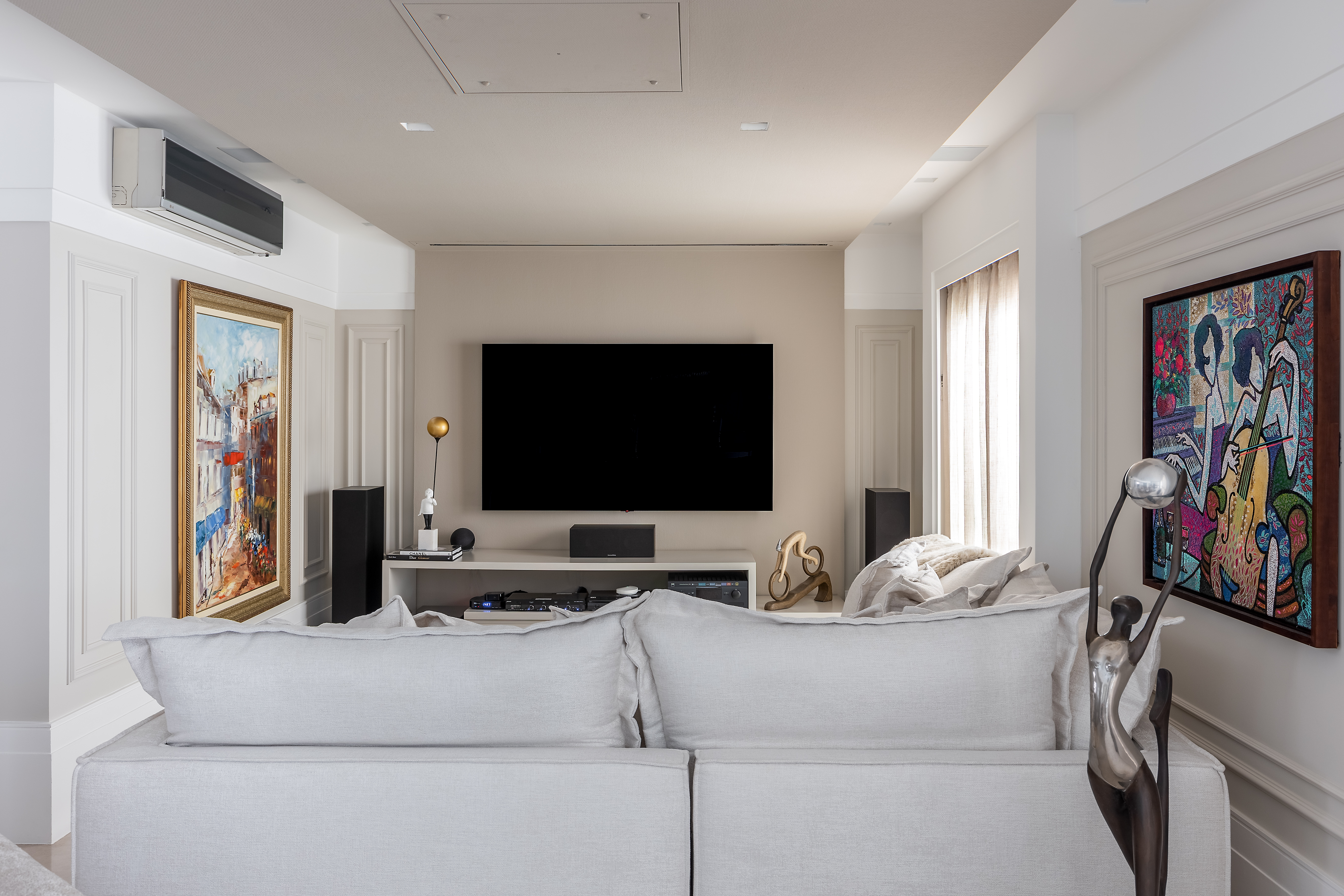 Sala de tv com sofá branco, rack branco e paredes com bosieries e quadros.