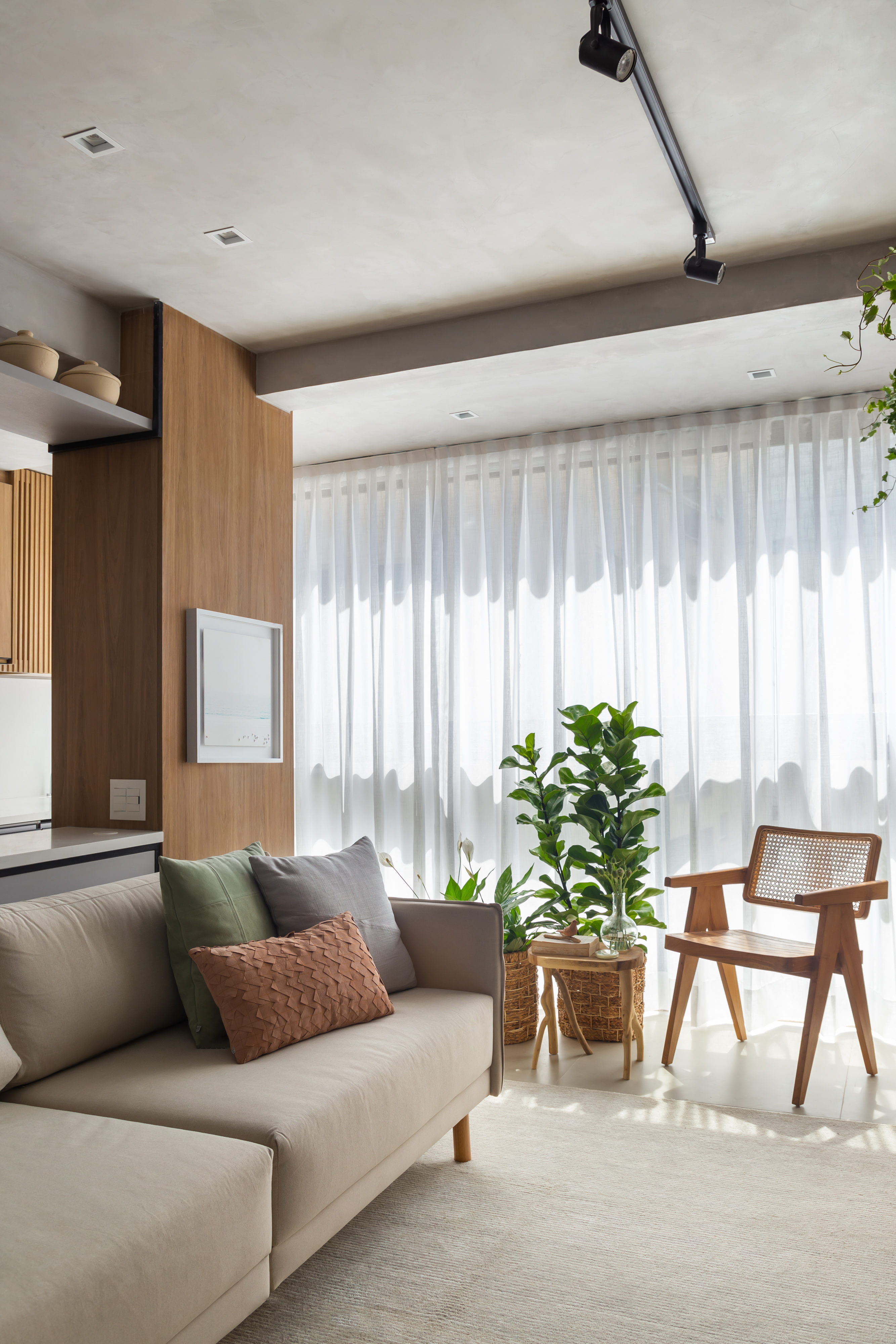 Sala de estar com sofá claro, tapete, cadeira, plantas e cortina branca.