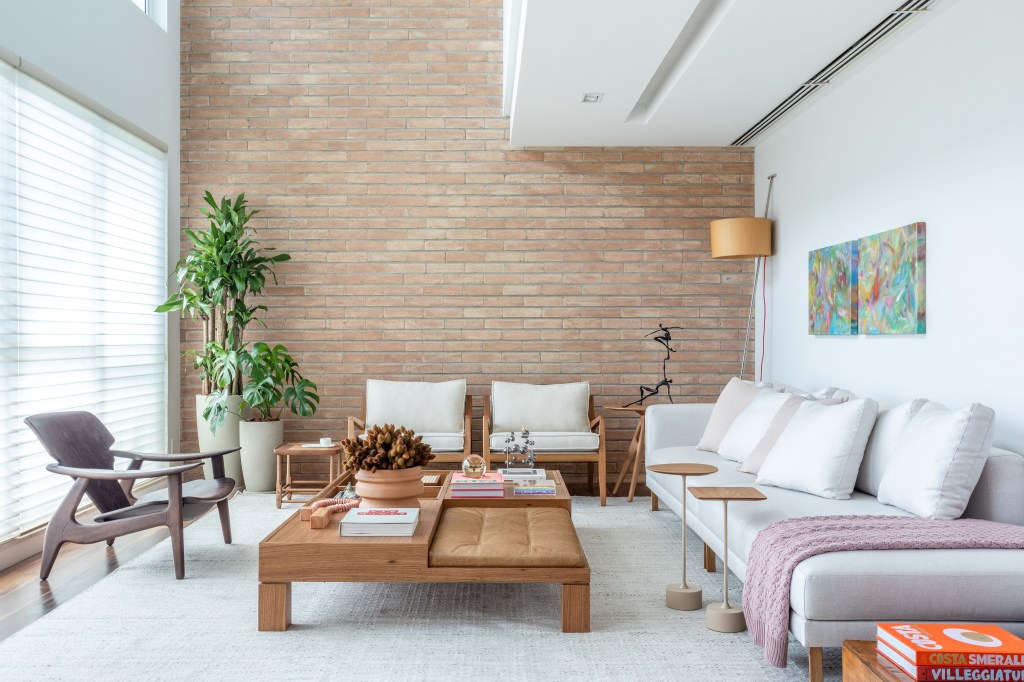 Sala de estar com pé-direito alto, parede de tijolinhos, sofá branco, poltronas e plantas.