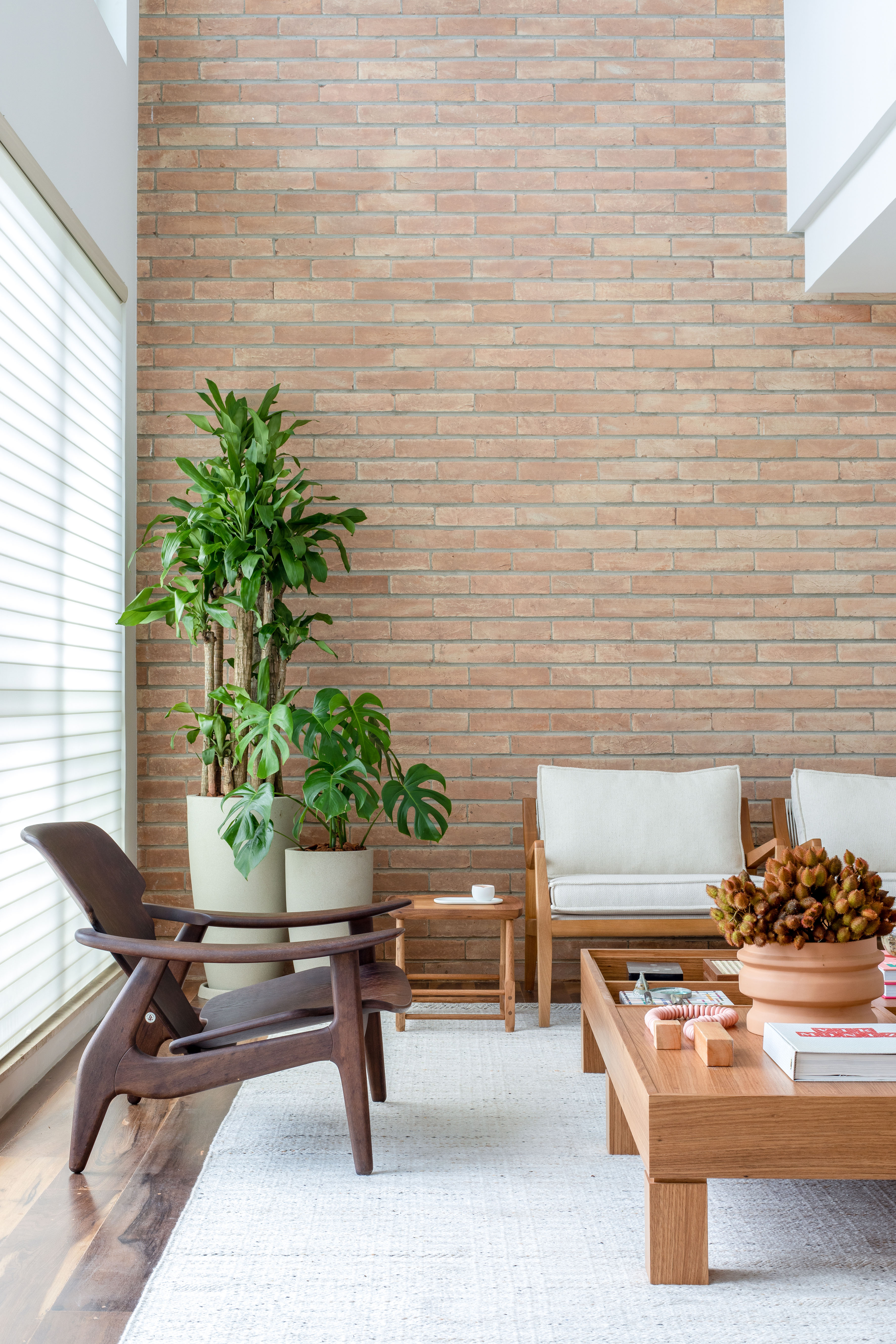 Sala de estar com pé-direito alto, parede de tijolinhos, sofá branco, poltronas e plantas.
