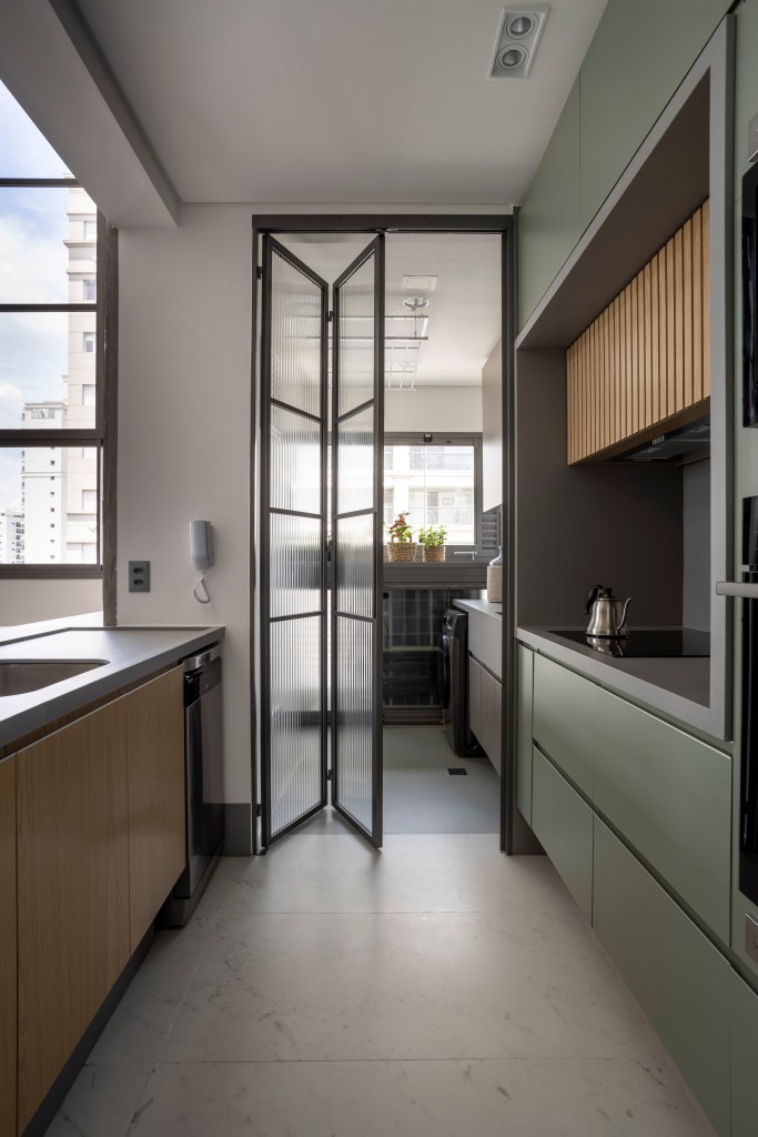Apê de 110 m² com pé-direito duplo vira duplex com mezanino metálico. NA foto cozinha separada da lavanderia por uma porta camarao de vidro canelado.