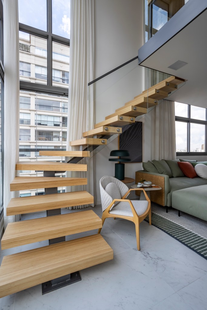 Apê de 110 m² com pé-direito duplo vira duplex com mezanino metálico. Na foto, escada de madeira leva ao mezanino metálico que serve de escritório;