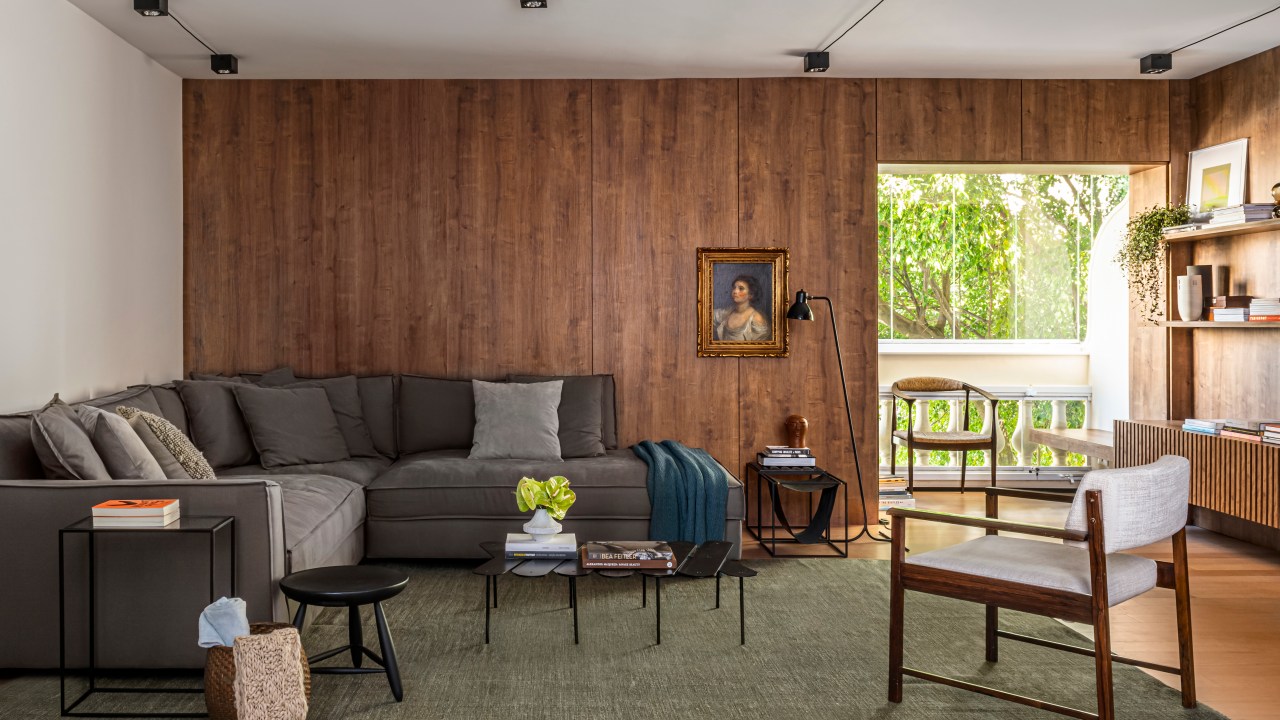 Sala de estar com parede revestida de madeira escura, tapete verde, sofá cinza escuro e luminária de piso.