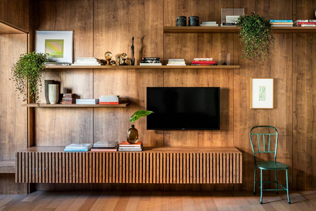 Sala de tv com parede revestida de madeira escura, rack com ripas e prateleiras com livros.