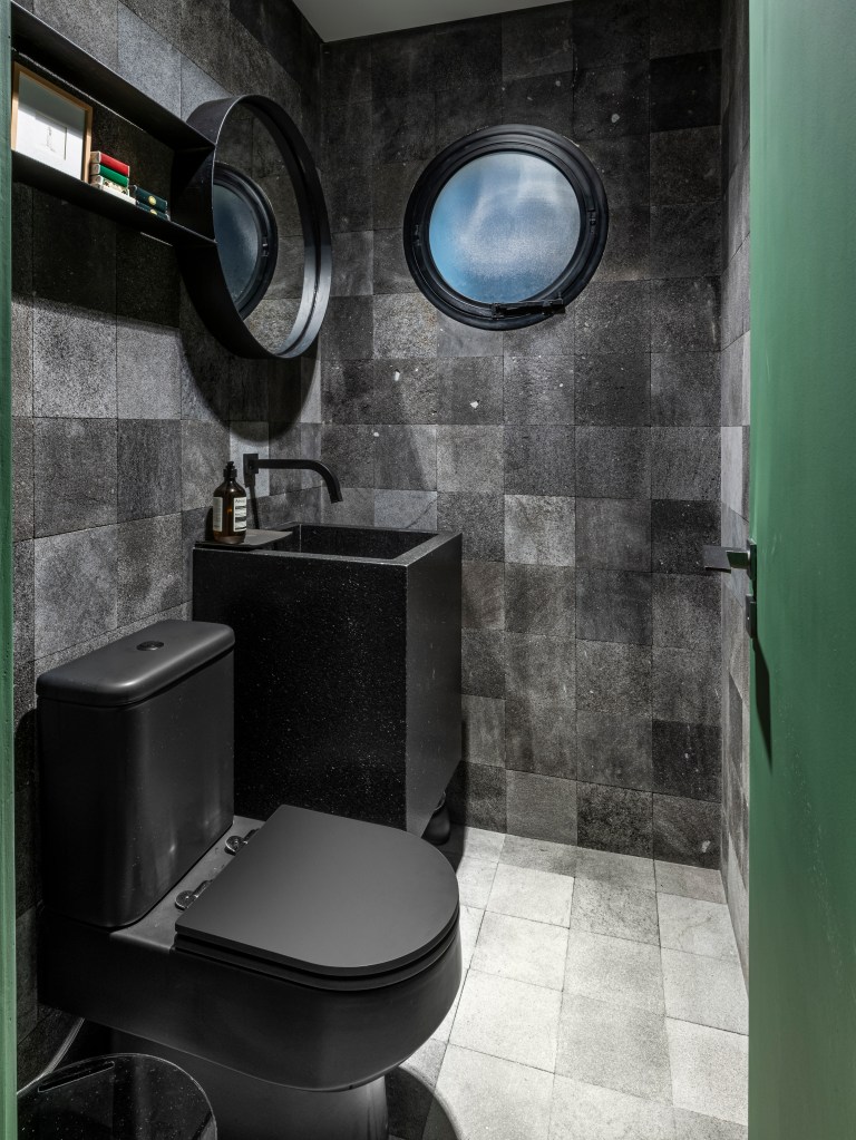 Banheiro com revestimento cinza, cuba preta esculpida, vaso sanitário preto e janela redonda.