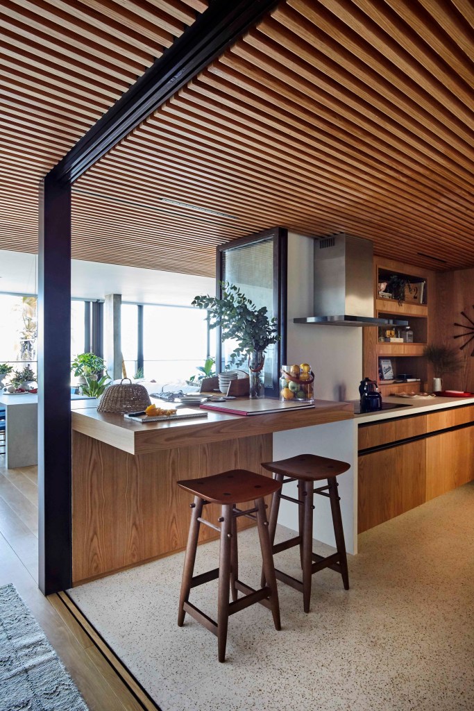 Cozinha integrada com revestimentos de madeira, teto de madeira ripada, bancada de madeira com duas banquetas.