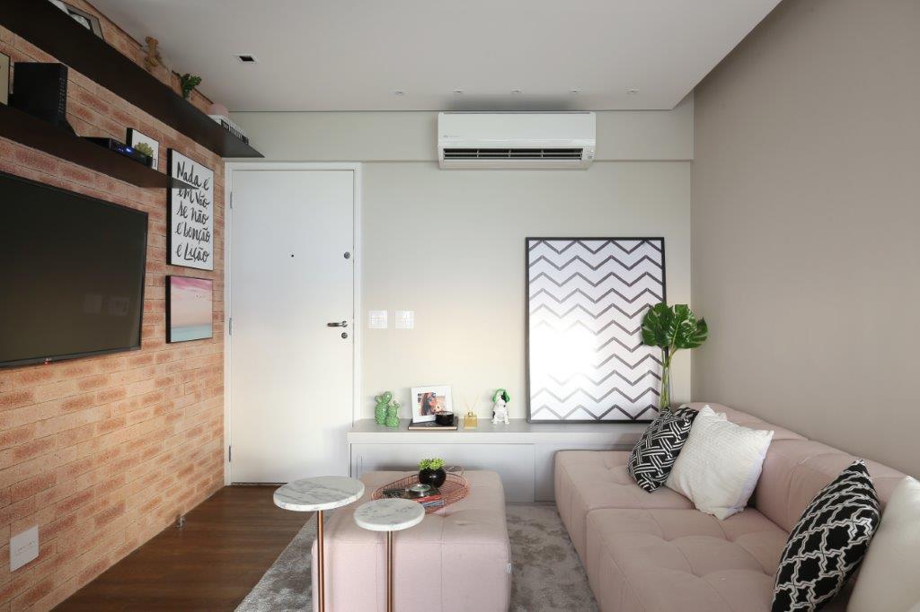 Sala de estar pequena com parede de tijolinho, tapete felpudo, sofá claro, pufe rosa.