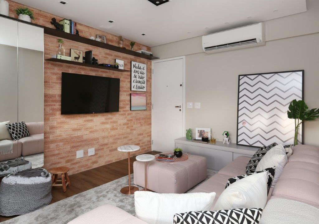 Sala de estar pequena com parede de tijolinho, tapete felpudo, sofá claro, pufe rosa.