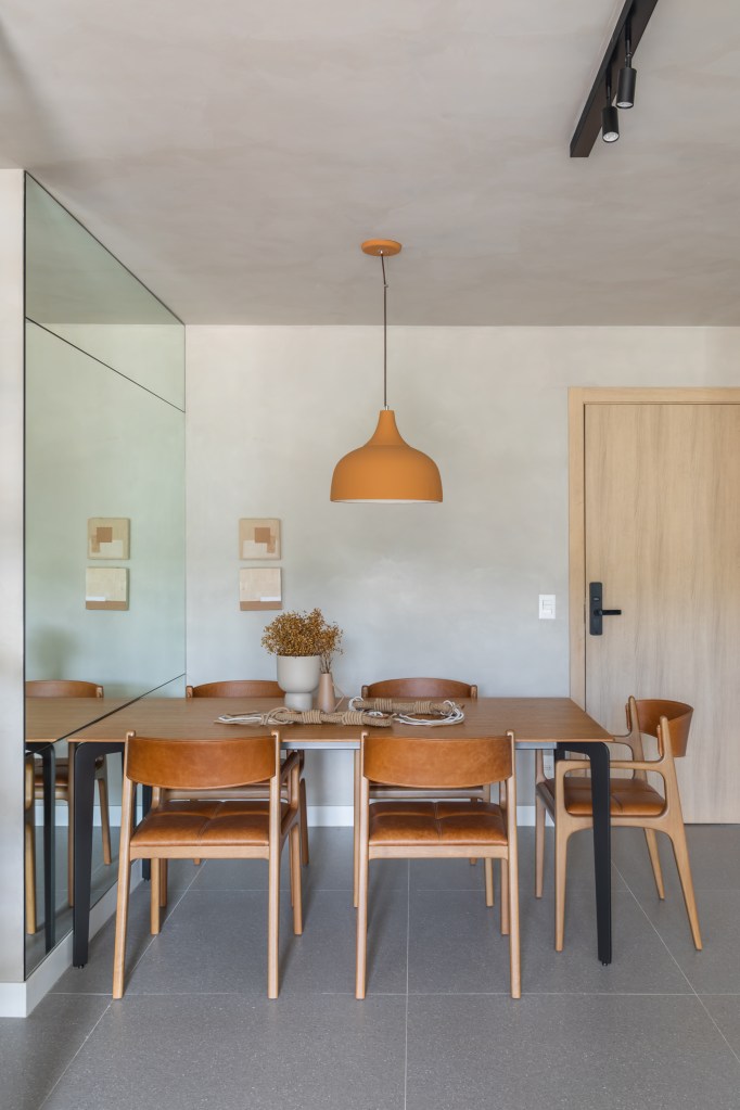 Sala de jantar com espelho na parede, mesa de madeira, cadeiras e luminária em tons terrosos.