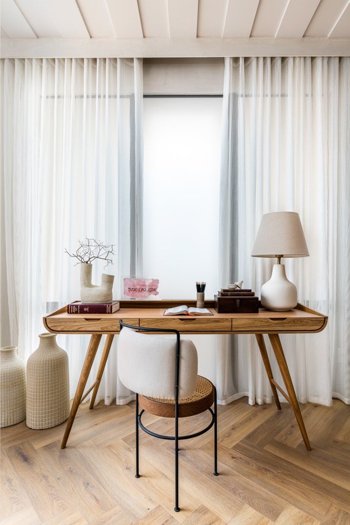 Home office pequeno claro com mesa de madeira, poltrona, abajur e cortina branca.