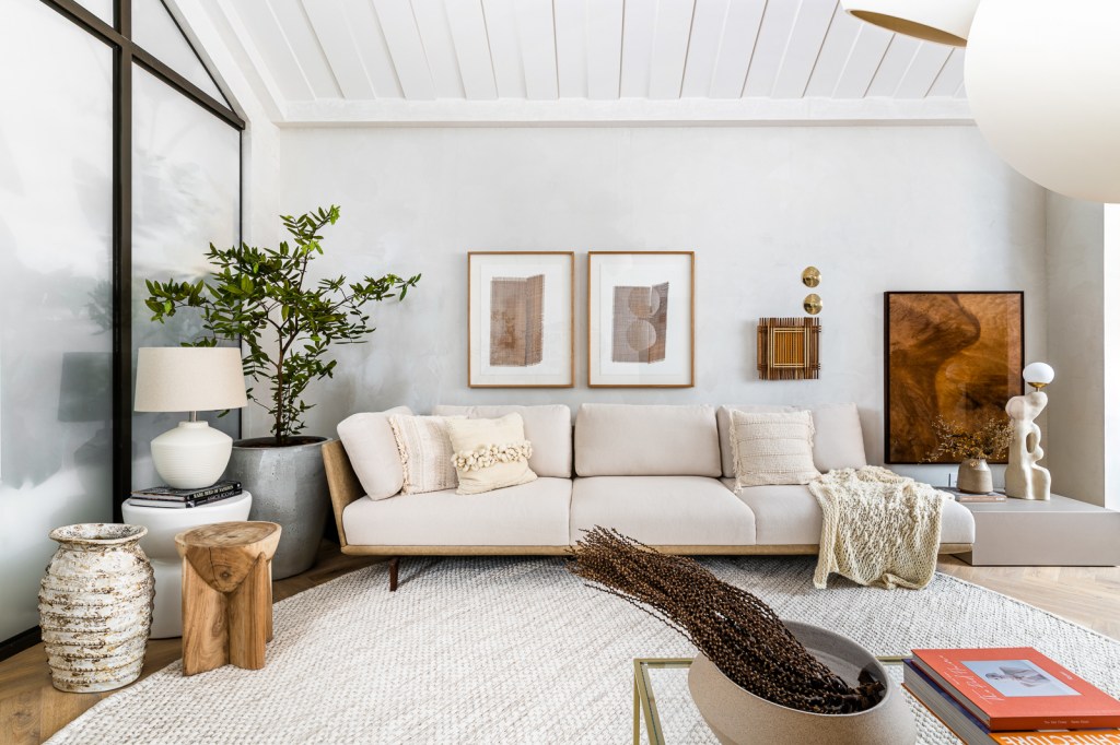 Sala de estar clara branca com tapete e sofá off white, planta e quadros em cima do sofá.