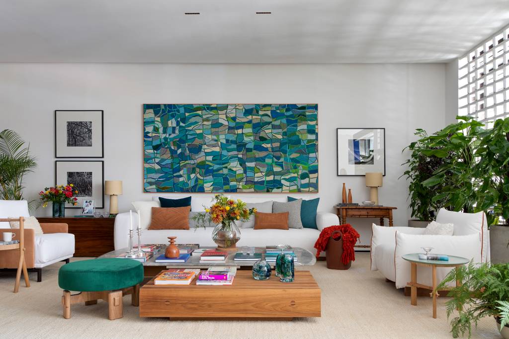 Casa que abraça: um projeto cozy, colorido e com clima carioca. Projeto de Migs Arquitetura na CASACOR SP 2023. Na foto, sala de estar com quadro bordado, sofá branco e vasos de plantas.