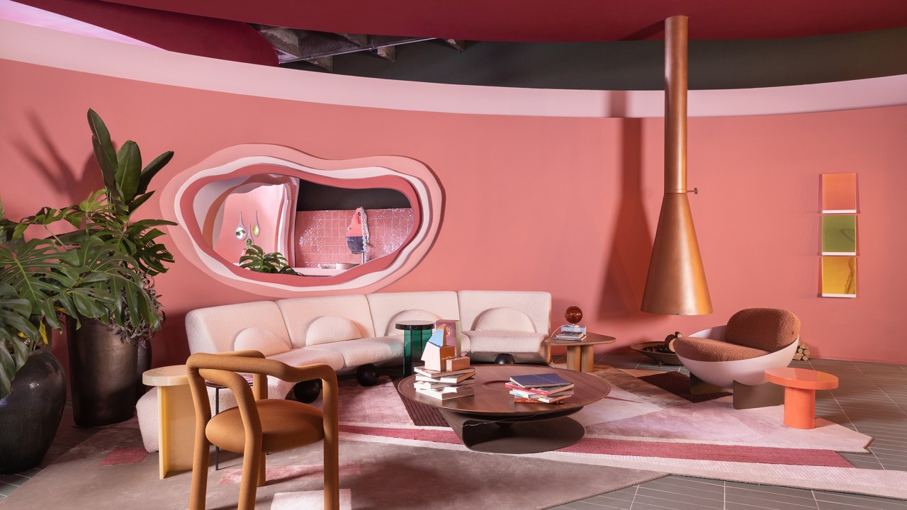 Casa rosa de 225 m² com cara de brinquedo feita para moradora de 64 anos. Projeto de Ricardo Abreu para a CASACOR SP 2023. Na foto, sala de estar com parede rosa, lareira e sofá curvo.