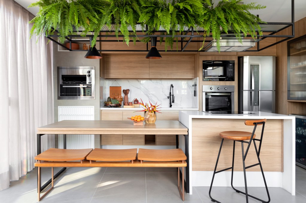 Cozinha gourmet integrada com prateleiras suspensa com plantas, bancada branca, banquetas, mesa de madeira. Sala com sofá ilha branco.