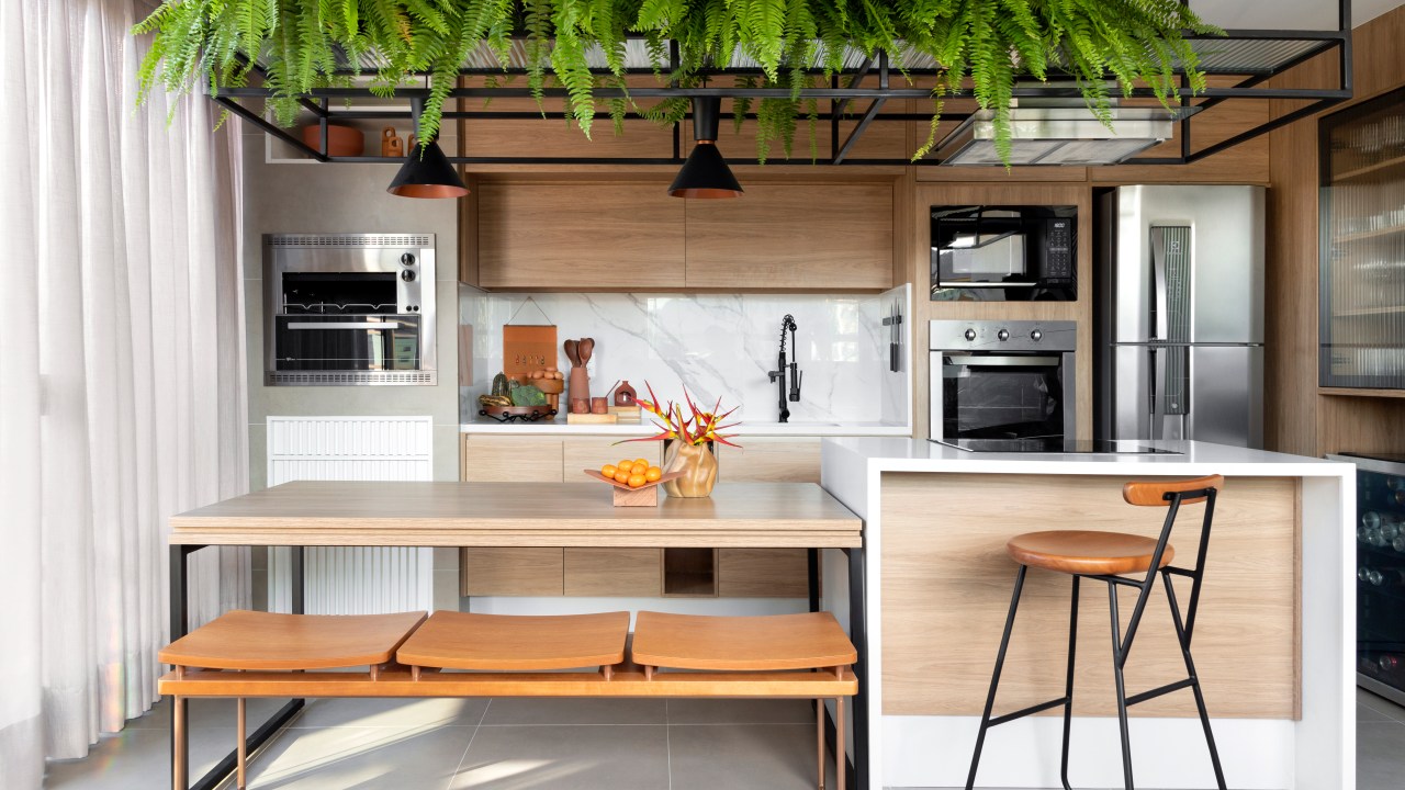 Cozinha gourmet integrada com prateleiras suspensa com plantas, bancada branca, banquetas, mesa de madeira. Sala com sofá ilha branco.