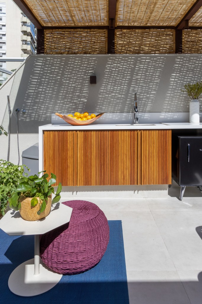 Terraço com bancada de cozinha, mesa lateral, tapete azul e pufe roxo.