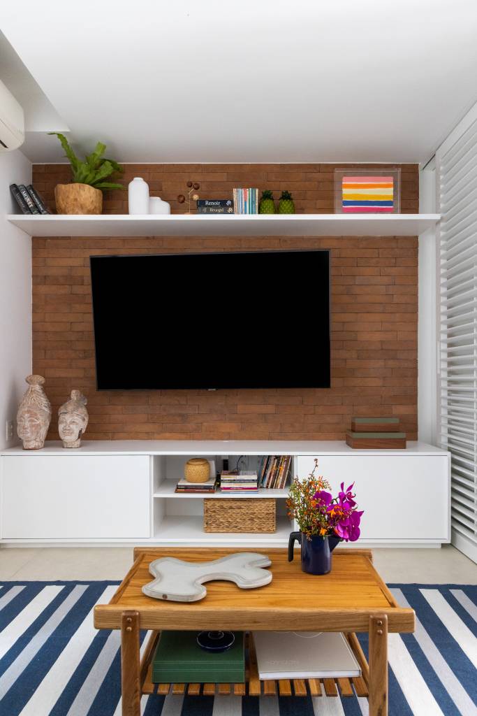 Sala de estar com tv, rack branco, tapete azul listrado, parede de tijolinhos.