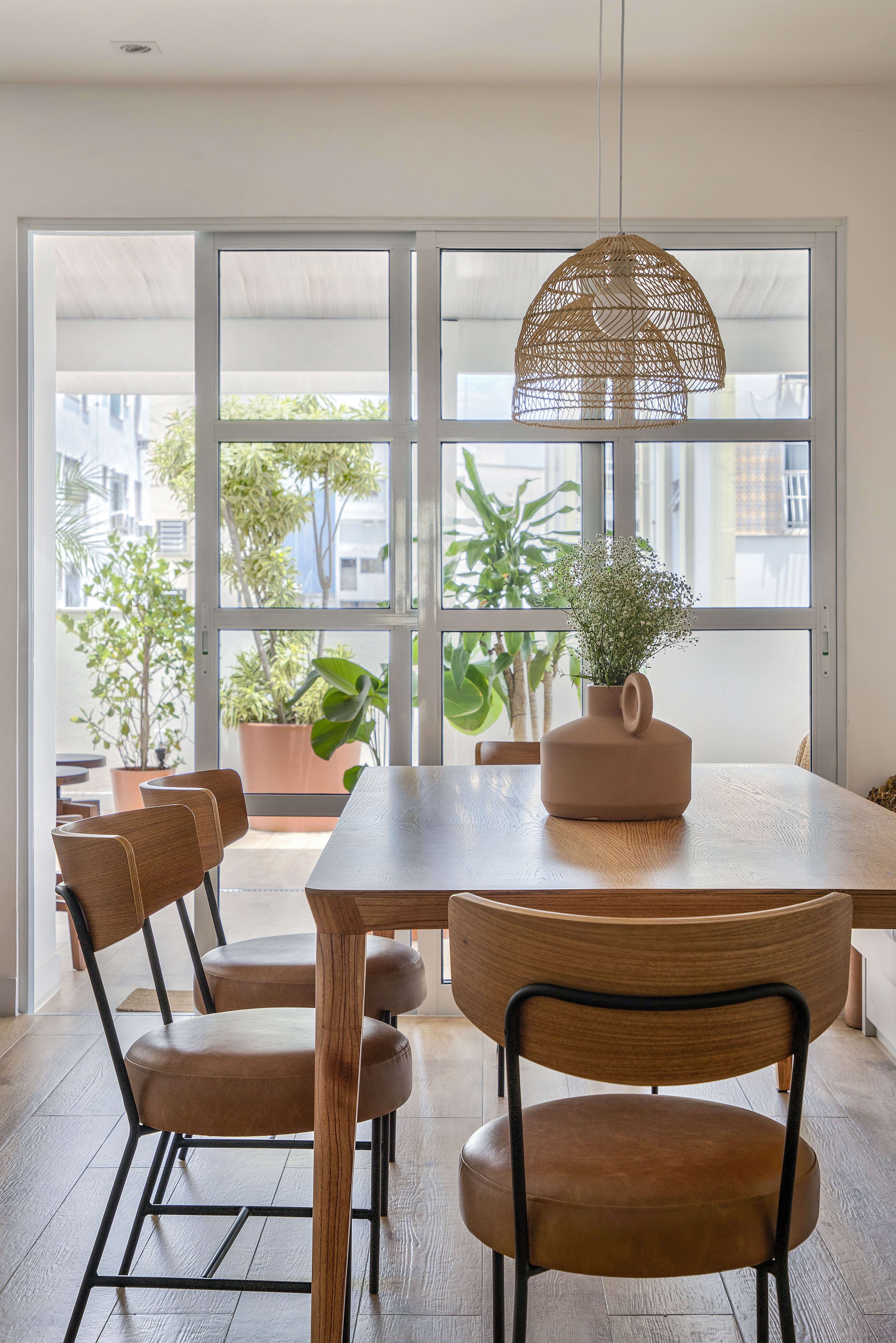 Sala de jantar branca clara com mesa de madeira, banco, luminária de palhinha e porta de vidro.