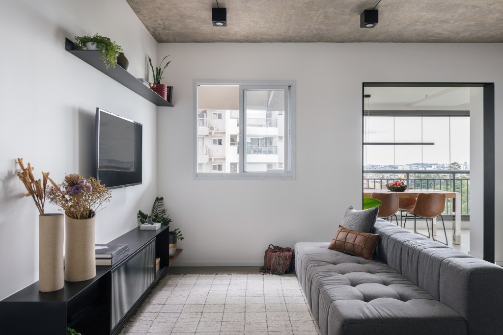 Sala de tv com sofá cinza e tapete claro, laje em concreto.