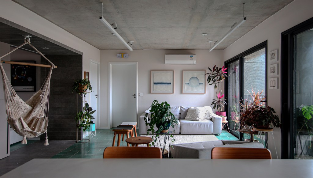Ladrilho hidráulico cria unidade visual neste apartamento de 140 m². Projeto do Studio MEMM. Na foto, sala de estar com varanda, cadeira suspensa e plantas.