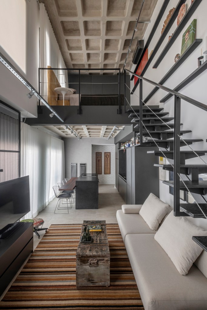 Loft dúplex com decoração industrial; sala com sofá branco, tapete listrado, cozinha ao fundo e escada preta.