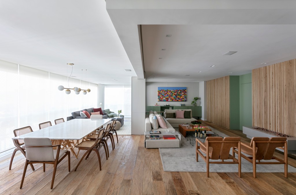Sala de estar integrada com jantar e varanda com piso de madeira, mesa de jantar branca e sala de estar com sofá claro.