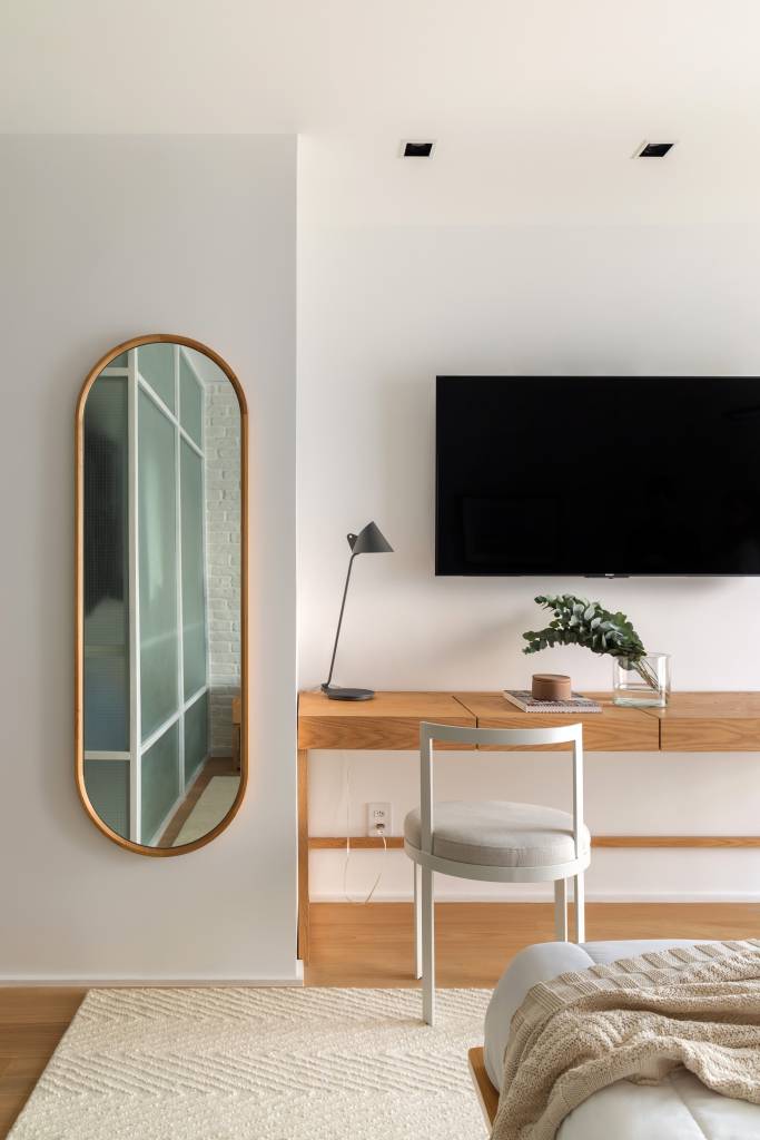 Quarto com espelho oval e home office com bancada de madeira e cadeira branca.