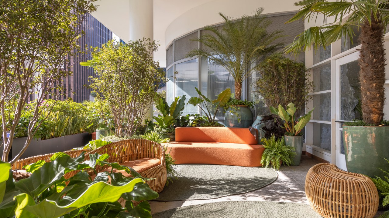 Jardim com vasos grandes, plantas, tapete verde e sofá curvo laranja.